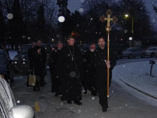 Clergy and faithful processing behind a cross through falling snow toward St. Andrew House